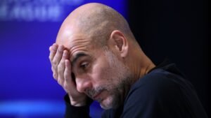 Pep Guardiola se couvrant le visage d'une main, affichant une expression sarcastique lors d'une conférence de presse concernant les dépenses de transfert de Manchester City.
