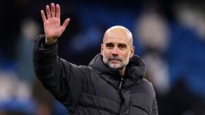 Pep Guardiola, vêtu d'un manteau noir, lève la main pour saluer, avec en arrière-plan flou un stade bleu, exprimant détermination et présence.