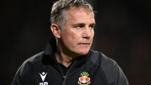 Phil Parkinson, Wrexham manager, looks pensive on the sidelines, wearing a black jacket with the club's logo visible.