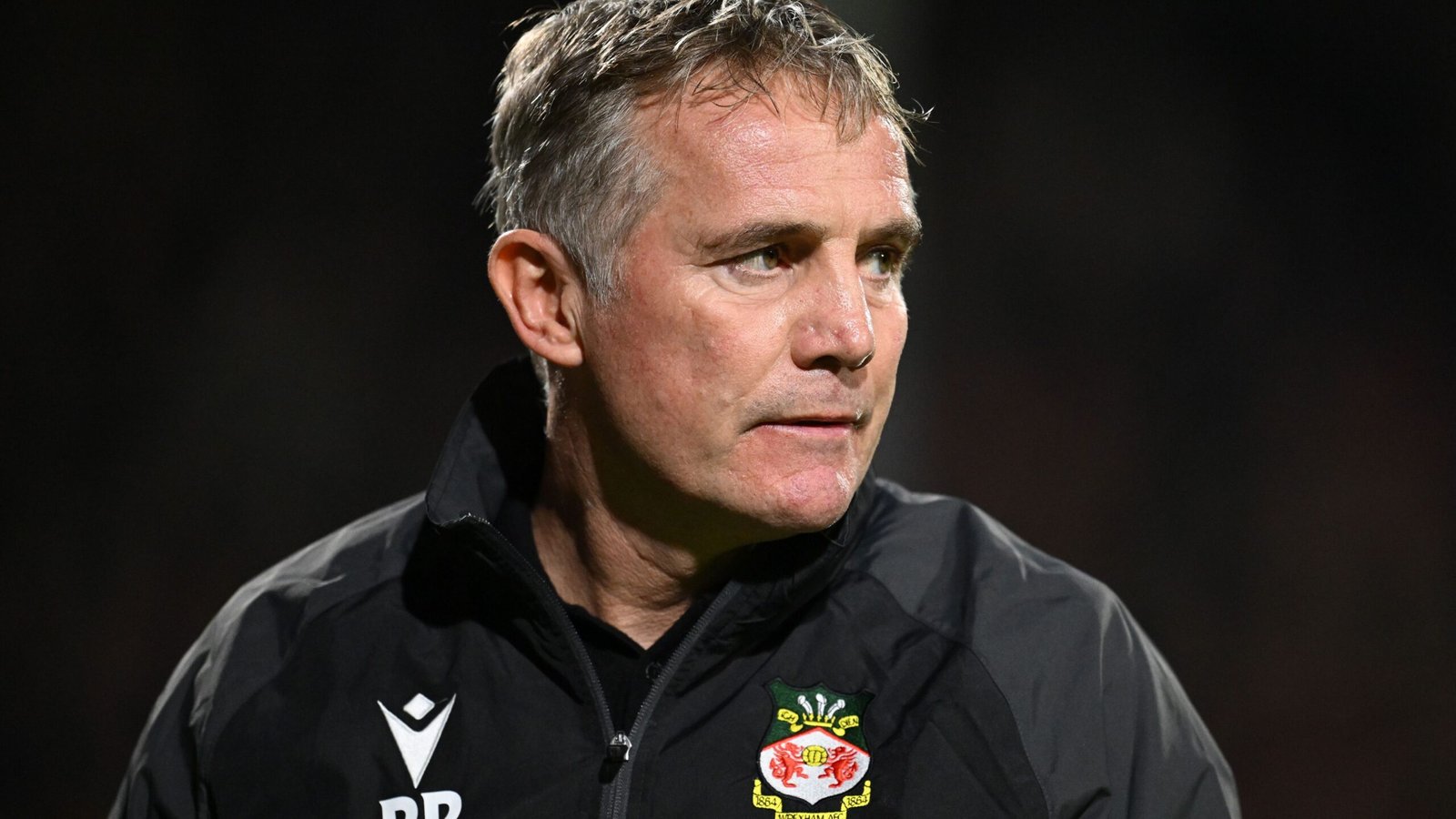 Phil Parkinson, Wrexham manager, looks pensive on the sidelines, wearing a black jacket with the club's logo visible.
