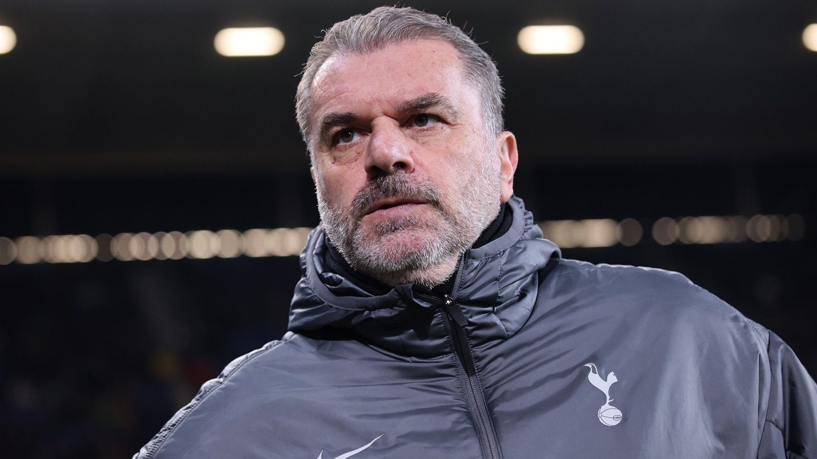 Ange Postecoglou, gekleed in een grijze Tottenham Hotspur-jas, kijkt serieus vanaf de zijlijn tijdens een wedstrijd.