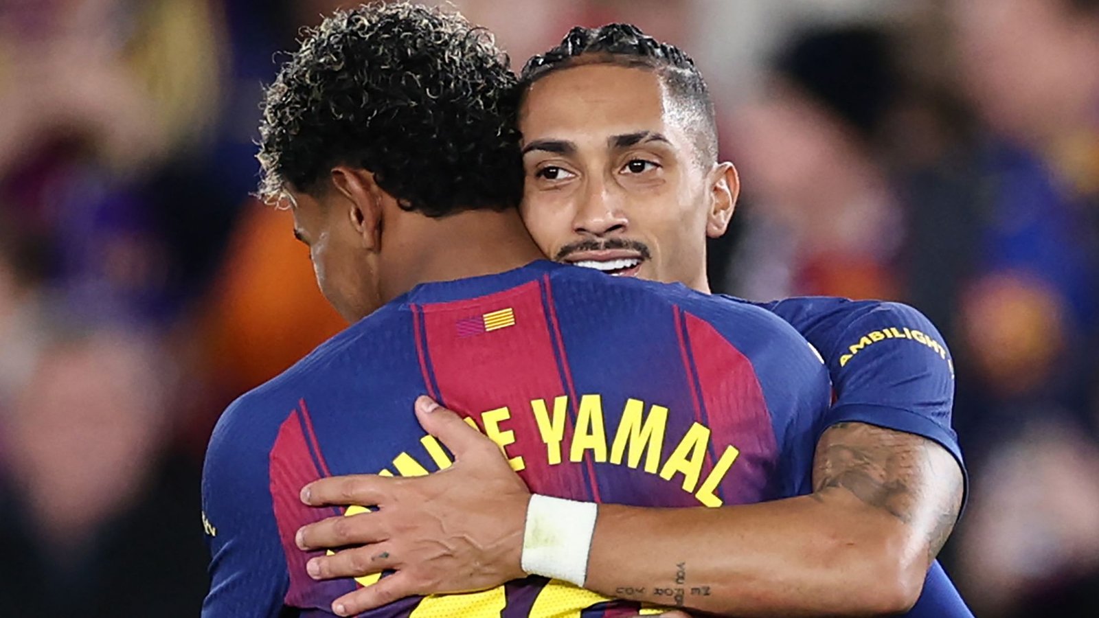 Raphinha embraces teammate Lamine Yamal after a match, both wearing Barcelona's distinctive claret and blue kit, celebrating their performance.