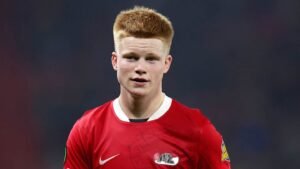 Young midfielder in a red club jersey with short blonde hair, showcasing concentration and determination during a match.