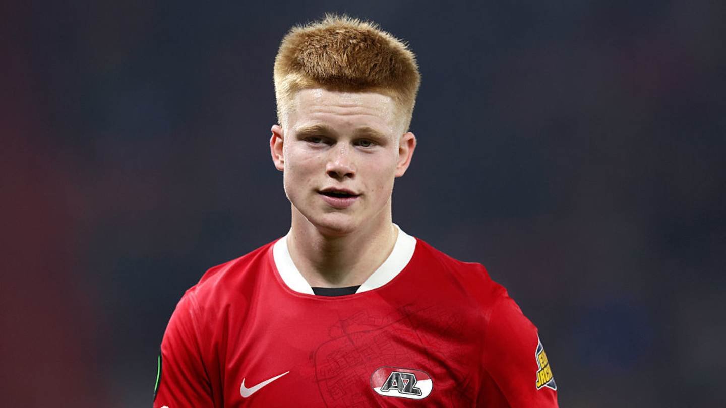 Young midfielder in a red club jersey with short blonde hair, showcasing concentration and determination during a match.