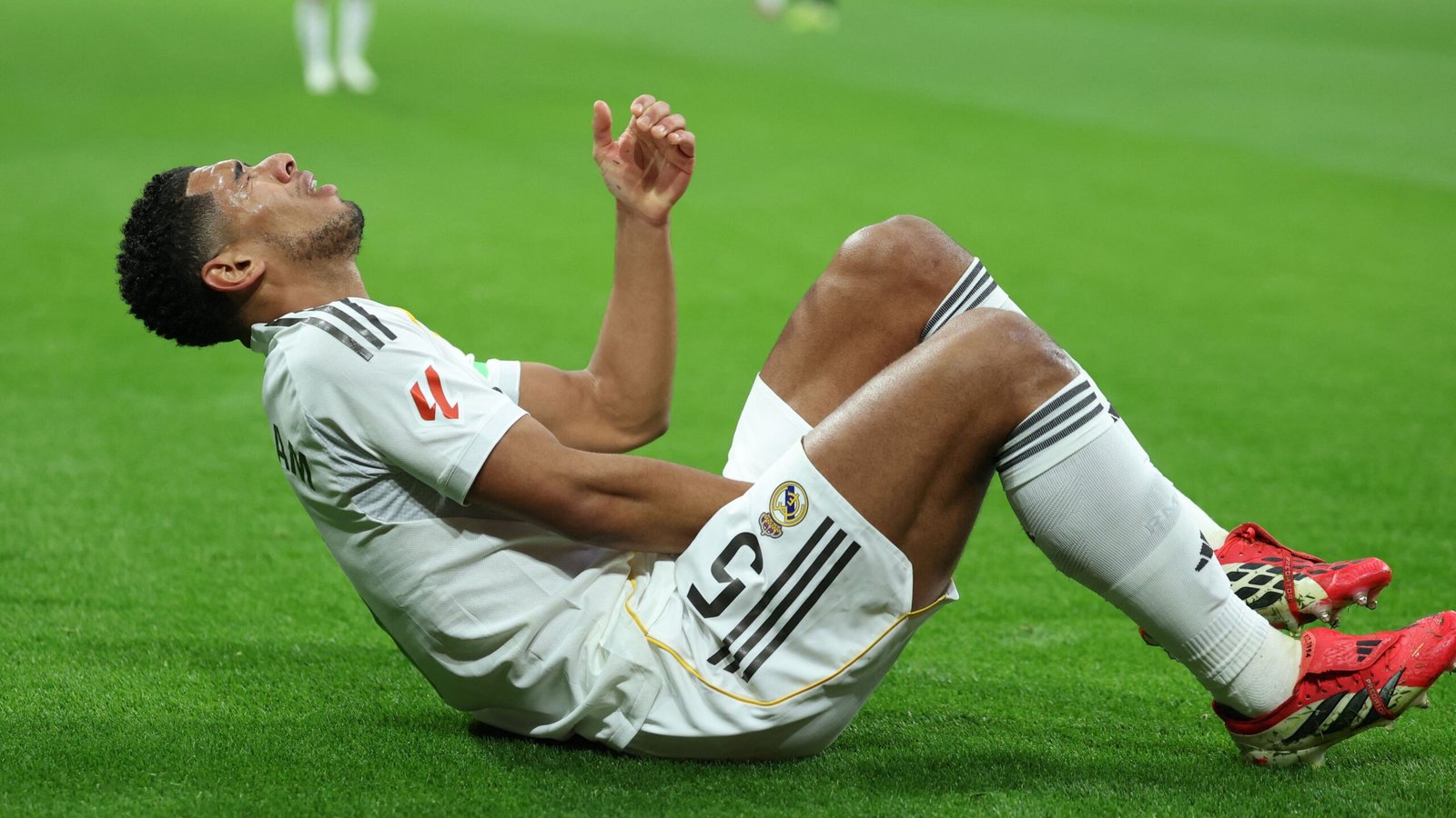 Jude Bellingham op het veld, die na een blessure tijdens een wedstrijd pijn uitdrukt, gekleed in het witte Real Madrid-tenue met rode schoenen.