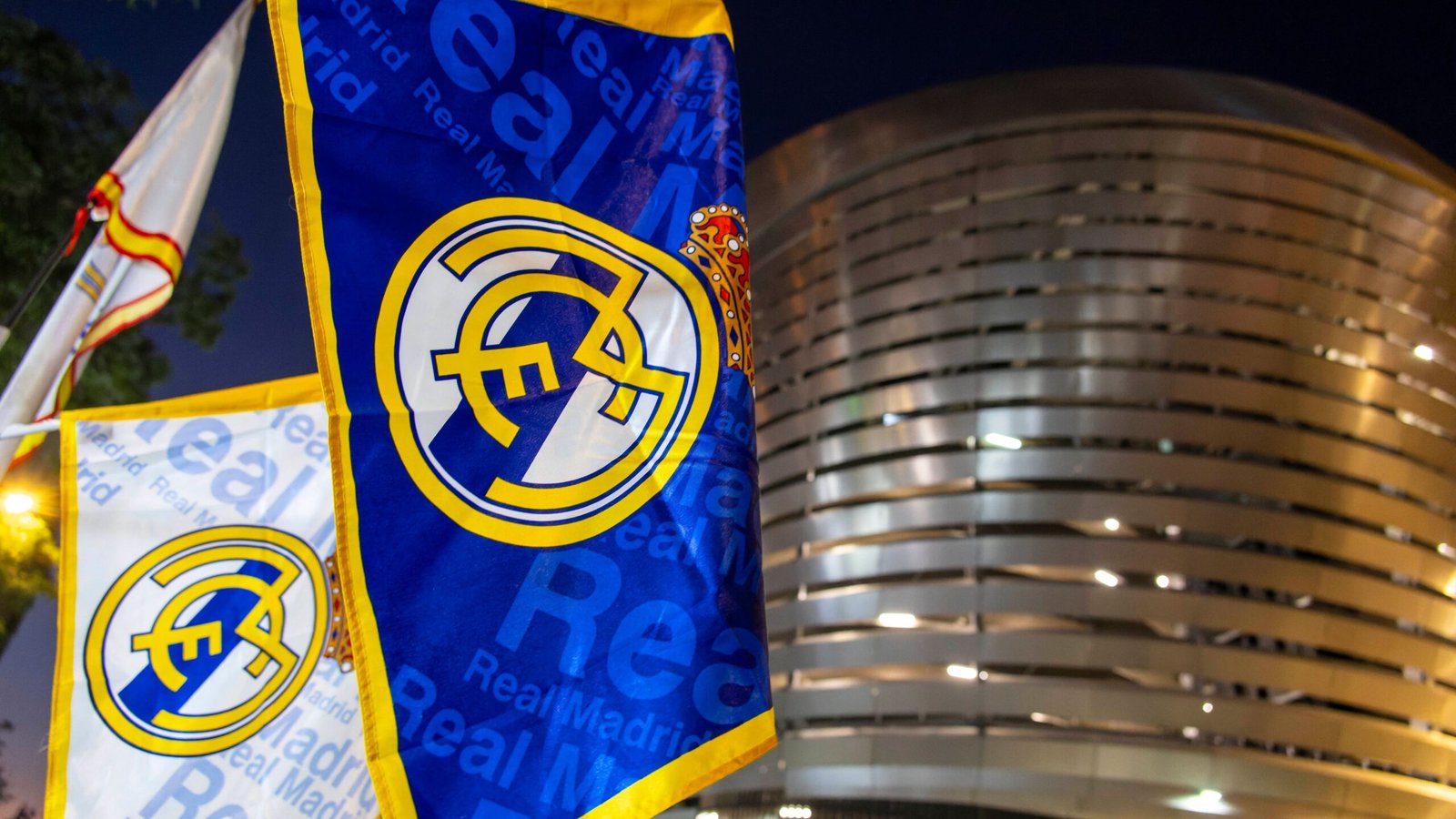 Des drapeaux du Real Madrid arborant l'écusson du club en bleu et jaune, se détachant sur fond du stade emblématique du Santiago Bernabéu de nuit.