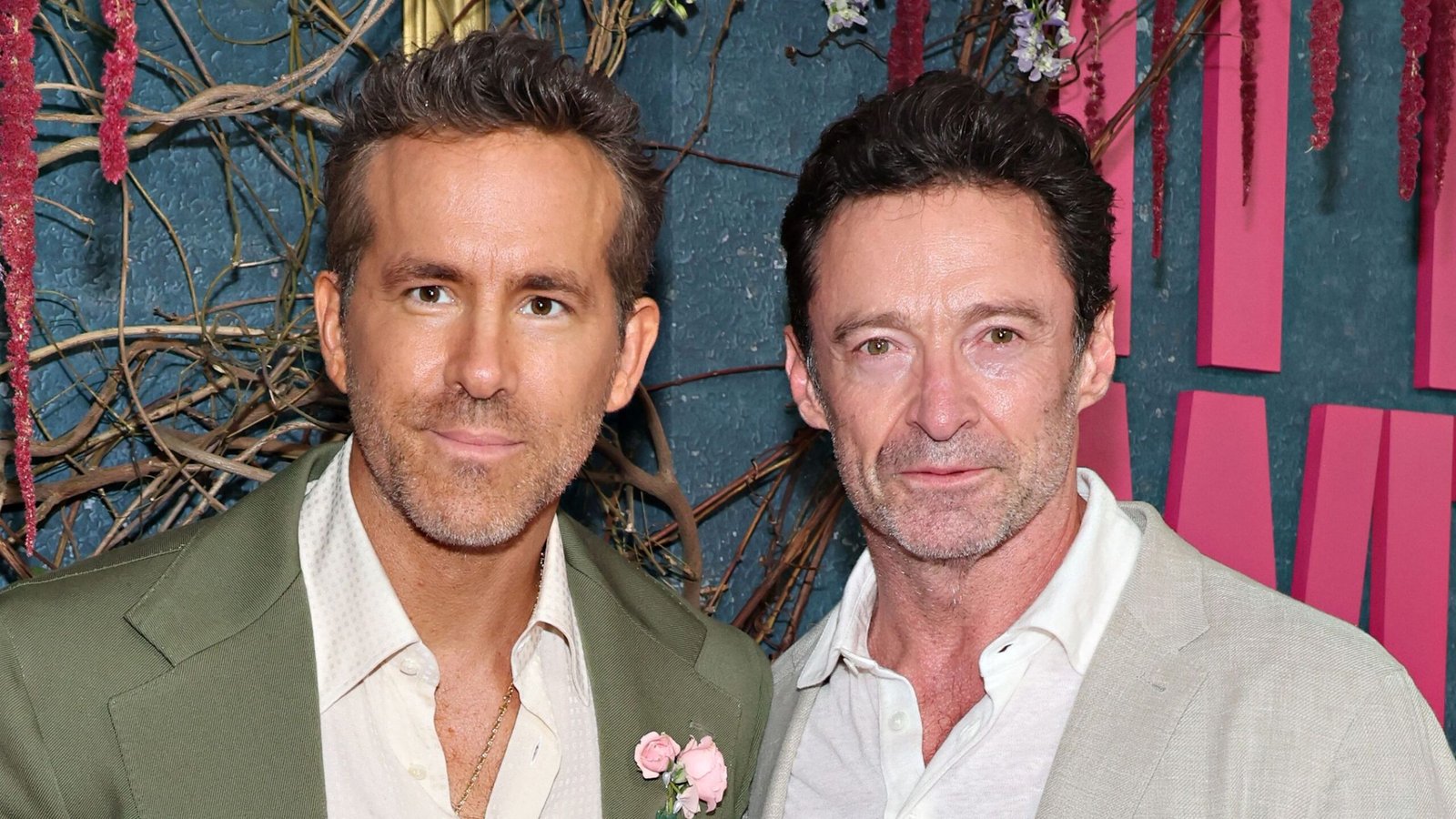 Ryan Reynolds et Hugh Jackman posent ensemble, tous deux vêtus de tenues élégantes, avec un fond à thème floral et des couleurs vibrantes.