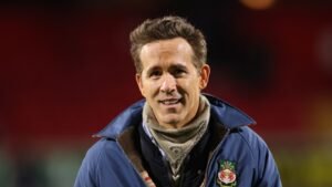Ryan Reynolds souriant chaleureusement dans un blouson du Wrexham AFC au stade, démontrant son soutien à l'équipe.
