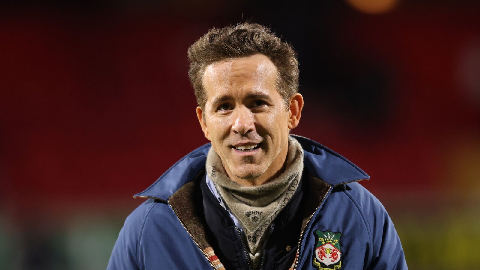 Ryan Reynolds souriant chaleureusement dans un blouson du Wrexham AFC au stade, démontrant son soutien à l'équipe.