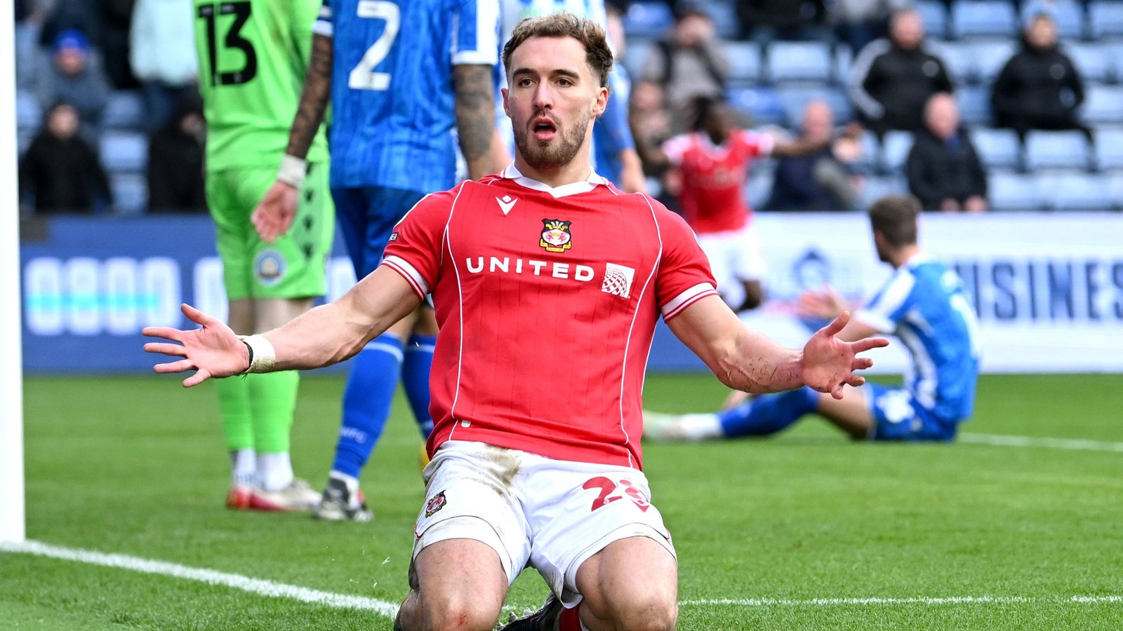 Wrexham-speler Sam Smith viert een doelpunt, gekleed in een rood shirt met "United" erop geborduurd, terwijl teamgenoten en tegenstanders op de achtergrond reageren.
