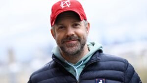 Jason Sudeikis, gekleed in een rode pet en een marineblauwe donsjas, glimlacht warmhartig in de buitenlucht, wat een ontspannen moment tijdens de opnames vastlegt.