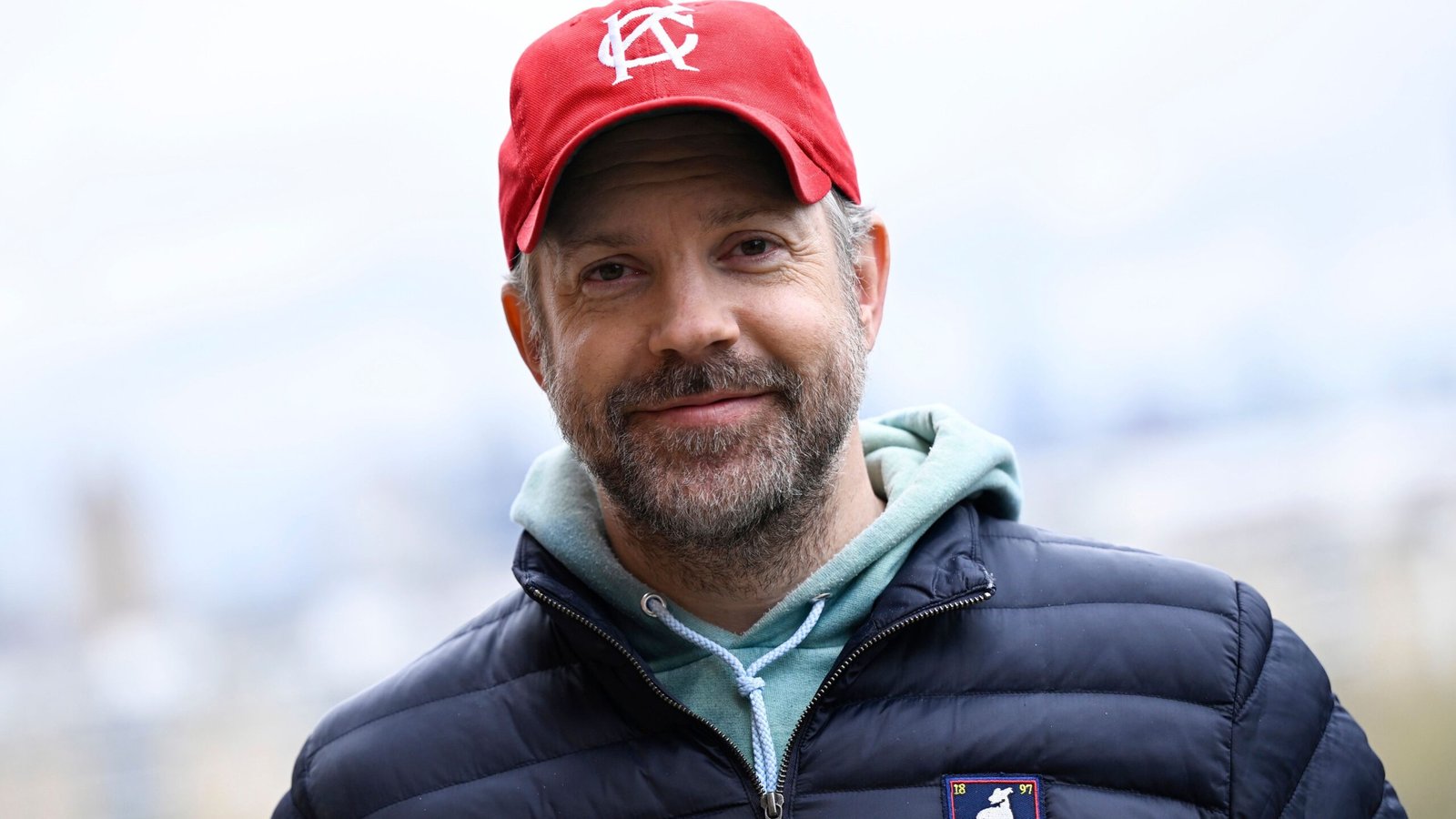 Jason Sudeikis, gekleed in een rode pet en een marineblauwe donsjas, glimlacht warmhartig in de buitenlucht, wat een ontspannen moment tijdens de opnames vastlegt.