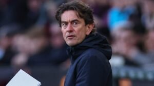 Thomas Frank, Brentford manager, focuses intently during a match, holding a notepad, with a blurred stadium backdrop.