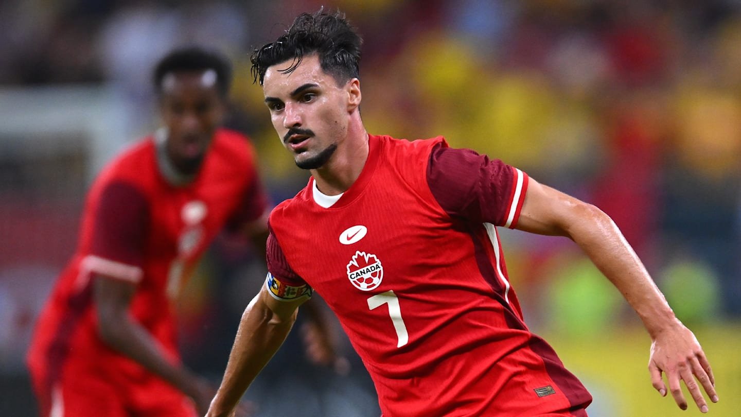 Canadese voetballer in een rood tenue met een esdoornblad, sprintend tijdens een wedstrijd, zijn vaardigheden en teamgeest tonend.