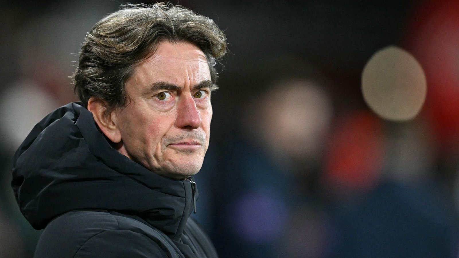 Thomas Frank, voormalig Liverpool-coach, kijkt bedachtzaam in een zwart jasje tijdens een wedstrijd, wat zijn serieuze houding benadrukt.