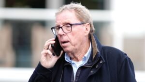 Harry Redknapp, voormalig Tottenham-manager, pratend aan de telefoon buitenshuis, gekleed in een donkere jas met een bril op.