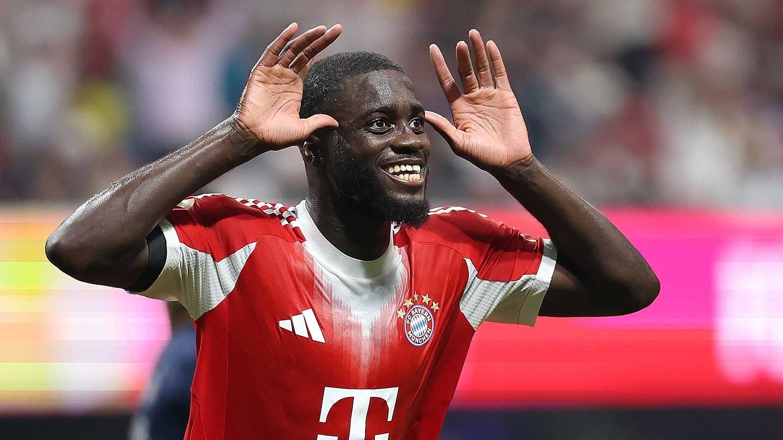 Bayern Munich player Dayot Upamecano celebrates joyfully, wearing the team's red and white kit with a background of cheering fans.