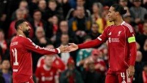 Mohamed Salah and Virgil van Dijk shake hands on the pitch during a Liverpool match, showcasing team camaraderie in the stands.