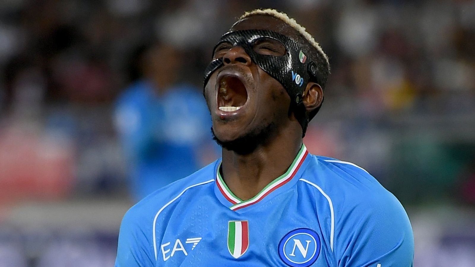 Victor Osimhen celebrating passionately in Napoli's blue kit, wearing a protective mask after scoring during a match.