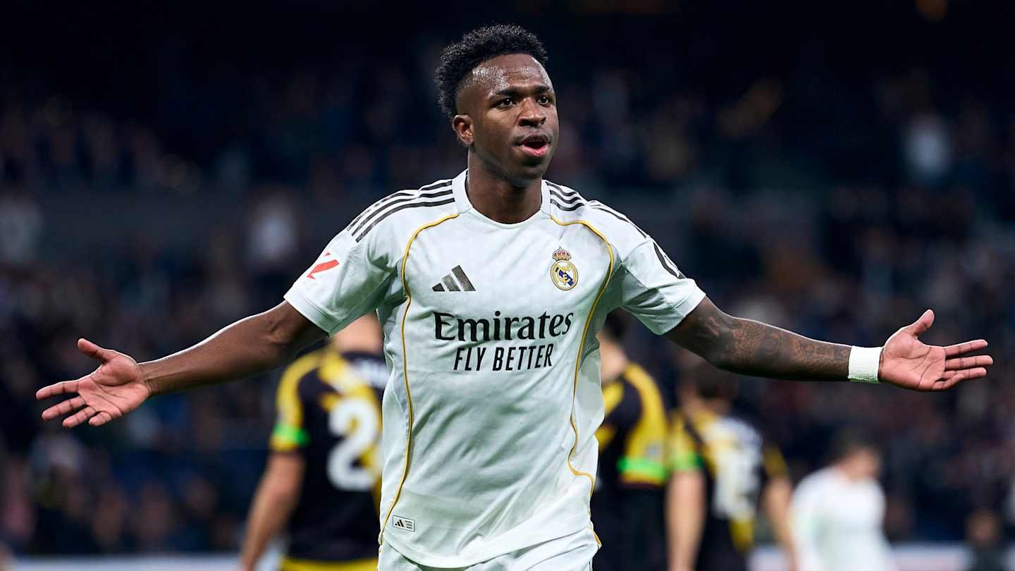 Vinícius Jr. celebrates a goal for Real Madrid, wearing the team's white kit with "Emirates" sponsor, amidst a crowd of players in the background.
