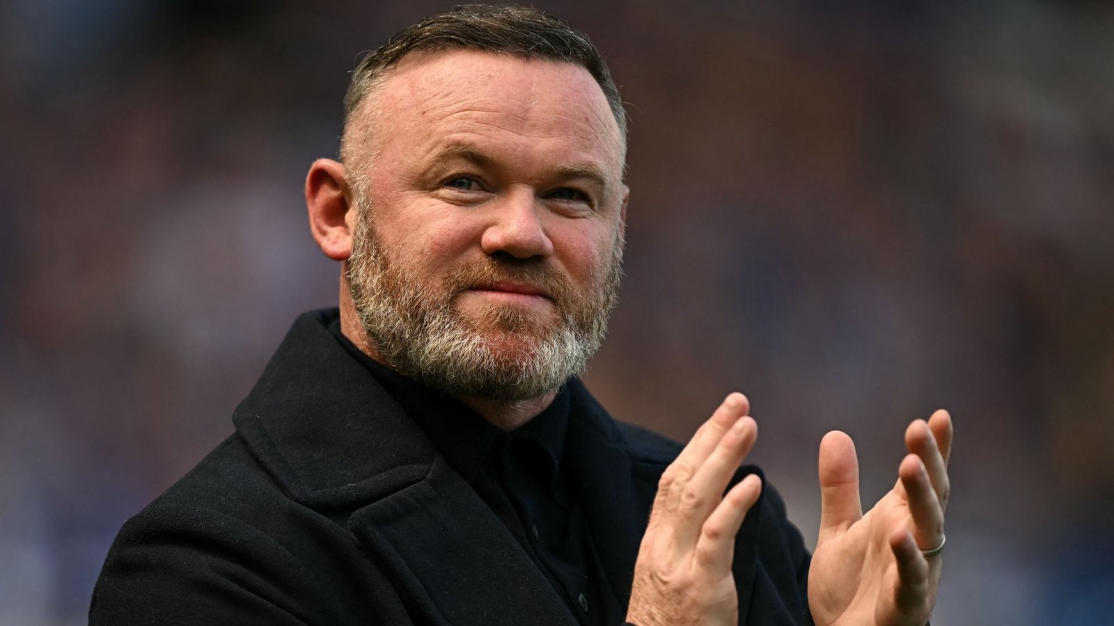 Wayne Rooney claps and smiles, dressed in a black coat, against a blurred stadium background, highlighting his role in football management.