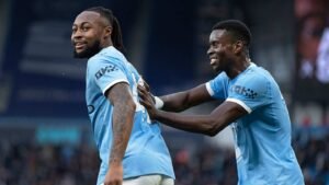 Deux joueurs de Manchester City célèbrent un but, l'un souriant et tirant taquinement le maillot de l'autre. Les deux portent la tenue bleu clair du club.