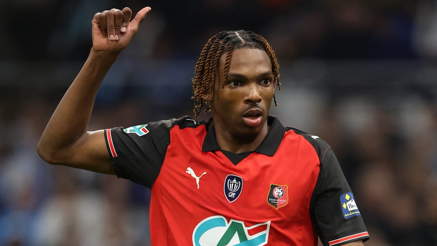 Jeremy Jacquet, in a red and black Rennes kit, gestures with his thumb up during a lively match, showcasing his enthusiasm.