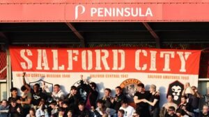 Salford City fans passionately singing beneath a large club banner, celebrating their team's history and camaraderie at a match.