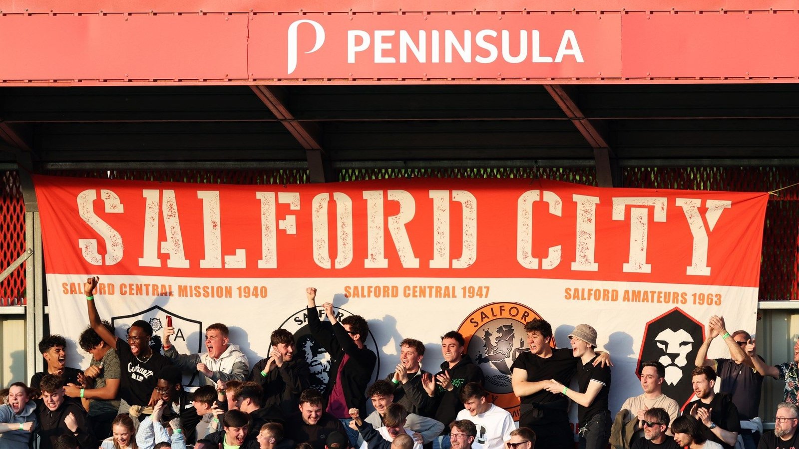 Salford City fans passionately singing beneath a large club banner, celebrating their team's history and camaraderie at a match.