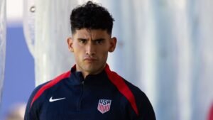 Un joueur de l'USMNT dans une veste d'entraînement marine avec des accents rouges, l'expression concentrée, se préparant pour un match ou une séance d'entraînement.