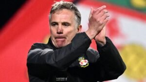 Phil Parkinson, Wrexham manager, applauds fans with a determined expression, set against Wrexham's flag colours in the background.