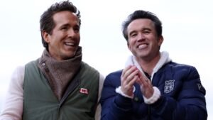 Ryan Reynolds and Rob McElhenney smiling and applauding during a Wrexham match, showcasing team spirit and excitement.