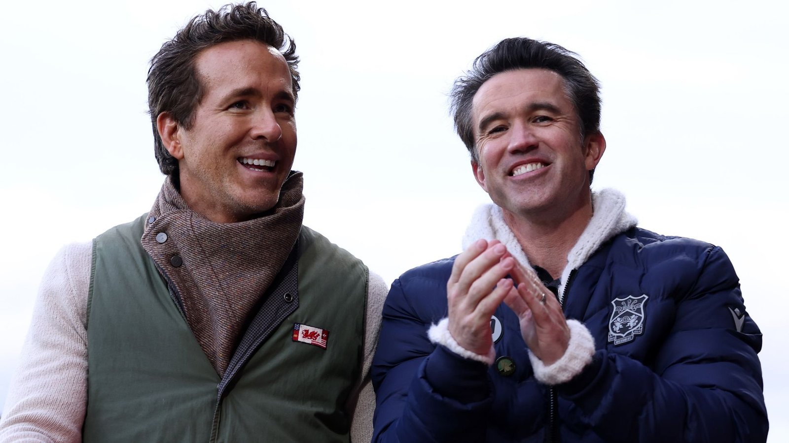 Ryan Reynolds and Rob McElhenney smiling and applauding during a Wrexham match, showcasing team spirit and excitement.