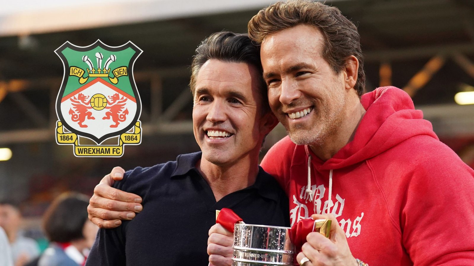 Ryan Reynolds et Rob McElhenney célèbrent en tenant un trophée, le logo du Wrexham FC apparaissant à leurs côtés sur fond de stade.