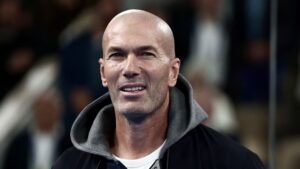 Zinedine Zidane souriant, portant une veste sombre avec une capuche grise, sur fond de stade flou, mettant en valeur sa présence charismatique.