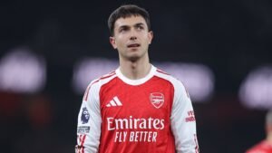 Zubimendi in Arsenal's red home kit, looking thoughtful during a match, showcasing Premier League branding and club sponsorship.