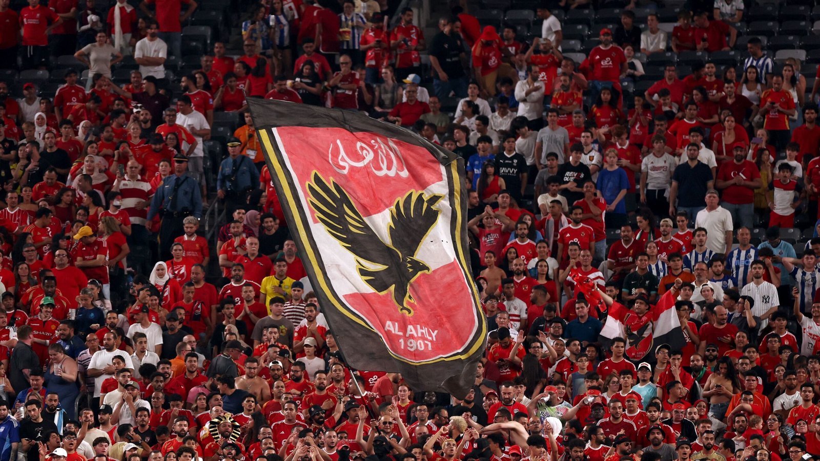 Al Ahly fans in vibrant red jerseys wave a large club flag featuring the eagle logo, creating an electrifying atmosphere in the stadium.