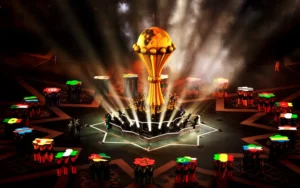 De gouden trofee van het Afrikaans Kampioenschap Voetbal, omgeven door verlichte tafels met de vlaggen van de deelnemende landen in een feestelijke sfeer.