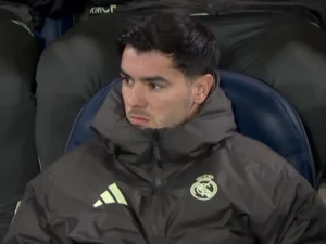 Real Madrid-speler zit op de bank, draagt een donker trainingsjack met het clubembleem, kijkt serieus tijdens een wedstrijd.