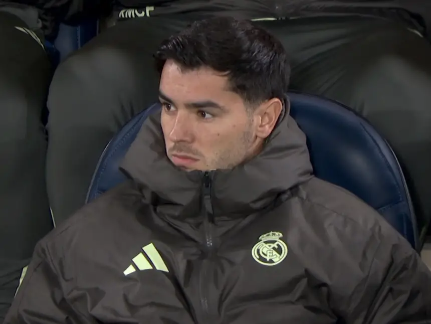 Real Madrid-speler zit op de bank, draagt een donker trainingsjack met het clubembleem, kijkt serieus tijdens een wedstrijd.