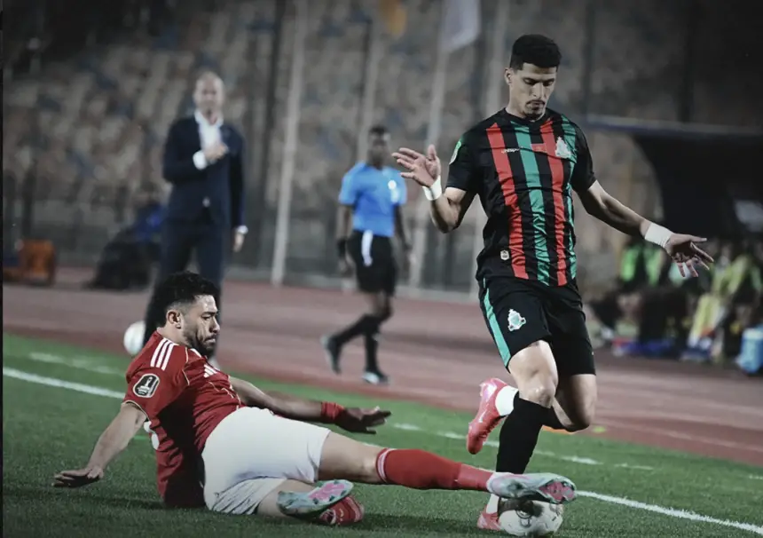 Moroccan player in green and black kit dribbles past opponent in red as fans and officials observe during a tense match action.