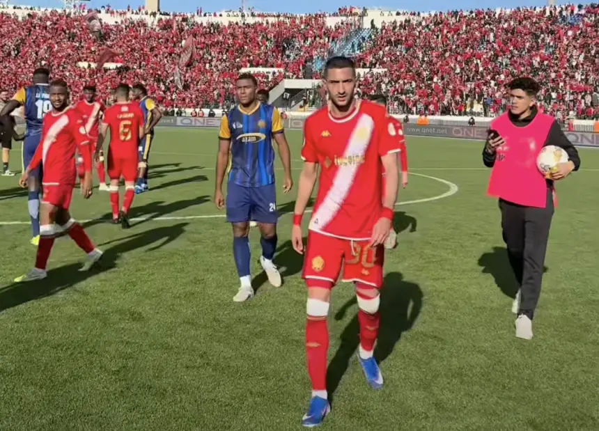 Celebrating players in red jerseys walking off the field, with a backdrop of enthusiastic fans clad in team colours.