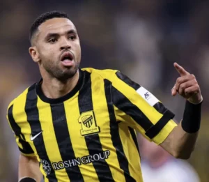 Ittihad Club player celebrating a goal, wearing a black and yellow striped kit, with an excited expression on the pitch.