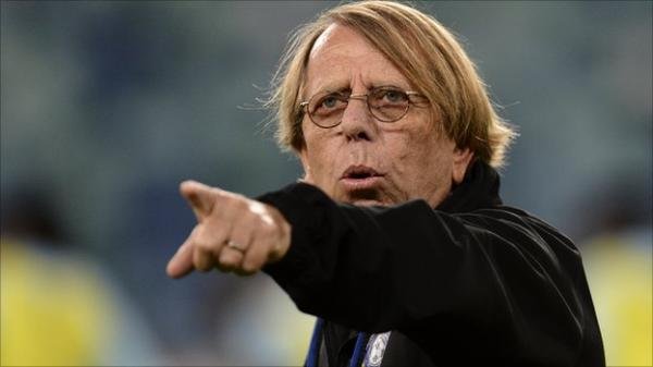 Claude Le Roy, Moroccan football coach, gestures passionately during a match, wearing a black jacket with glasses and tousled hair.