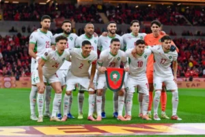 Het Marokkaans nationaal voetbalelftal poseert voor een teamfoto, gekleed in witte tenues met groene accenten en houdt een schild vast met daarop een leeuwenembleem.