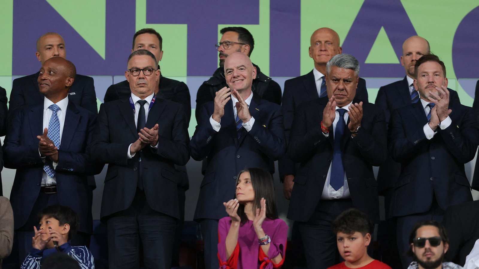 Voetbalfunctionarissen applaudisseren op de tribune, gekleed in donkere pakken, tijdens een belangrijke wedstrijd in een stadion, met fans zichtbaar op de voorgrond.