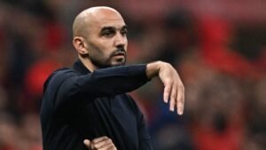 Walid Regragui gesturing on the sidelines during a match, wearing a black shirt, with a blurry background of fans in red.