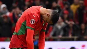 Moroccan player with head down in disappointment during a match, wearing the national team's red and green kit, with fans in the background.
