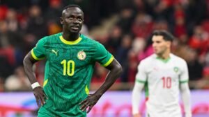 Sadio Mané in a green Senegal kit with yellow accents, looking frustrated; Moroccan player in white and green, background action from a match.