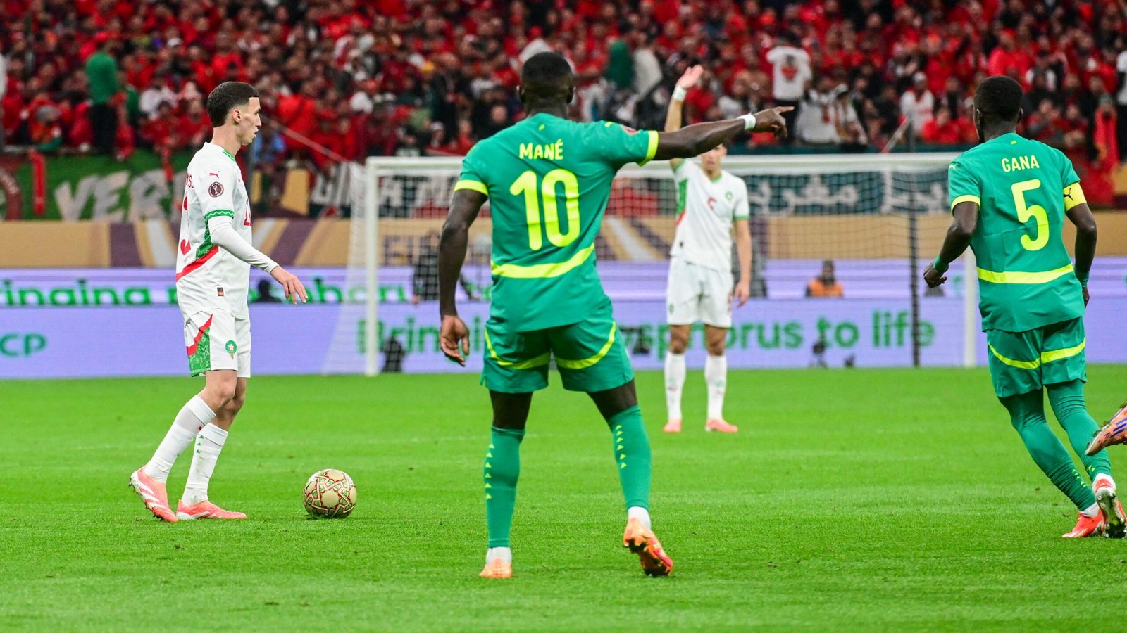 Marokkaanse speler in wit shirt op het veld terwijl Senegal's Sadio Mané in groen gebaart, te midden van een levendige menigte tijdens een gespannen AFCON-wedstrijd.